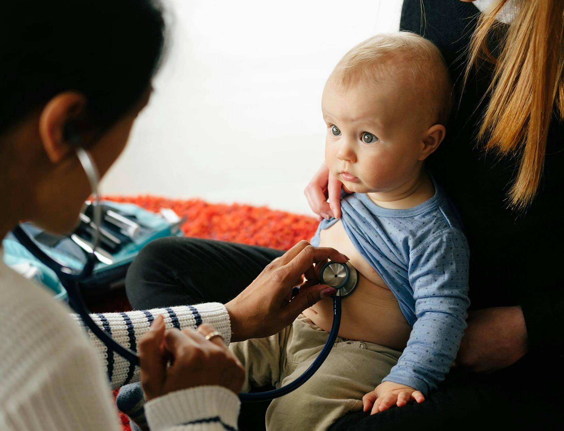Lege undersøker barn på mors fang med stetoskop, på hjemmebesøk